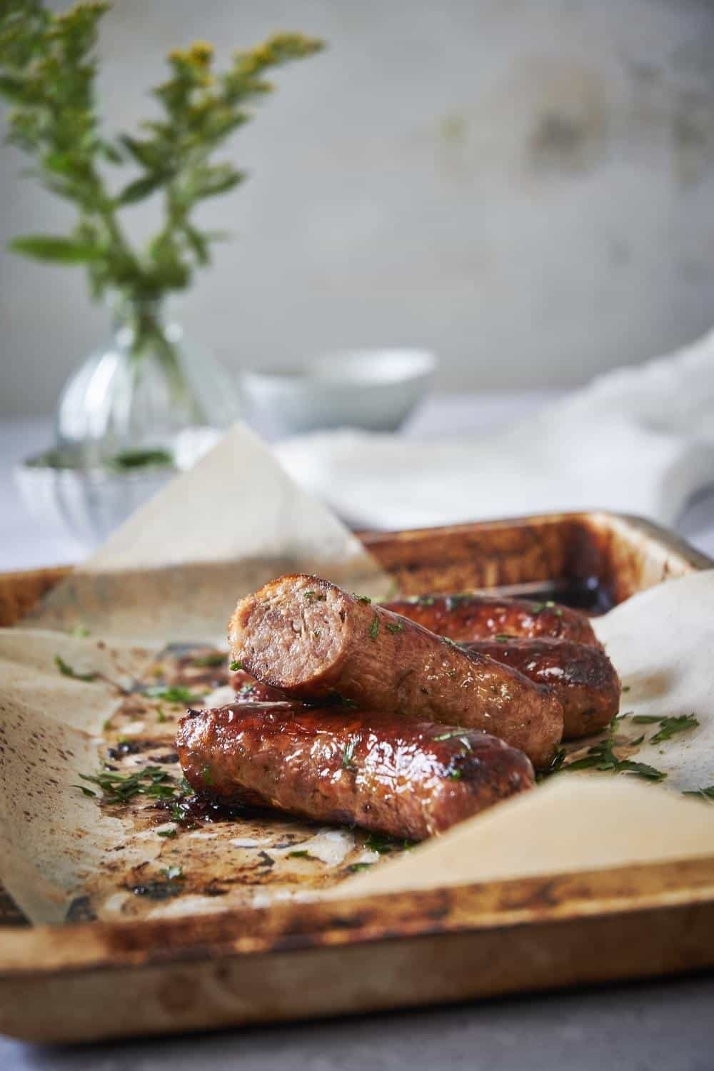 Four brats on a sheet pan lined with parchment paper garnished with parsley. One of the brats is cut in half to show the pink and juicy cross section.