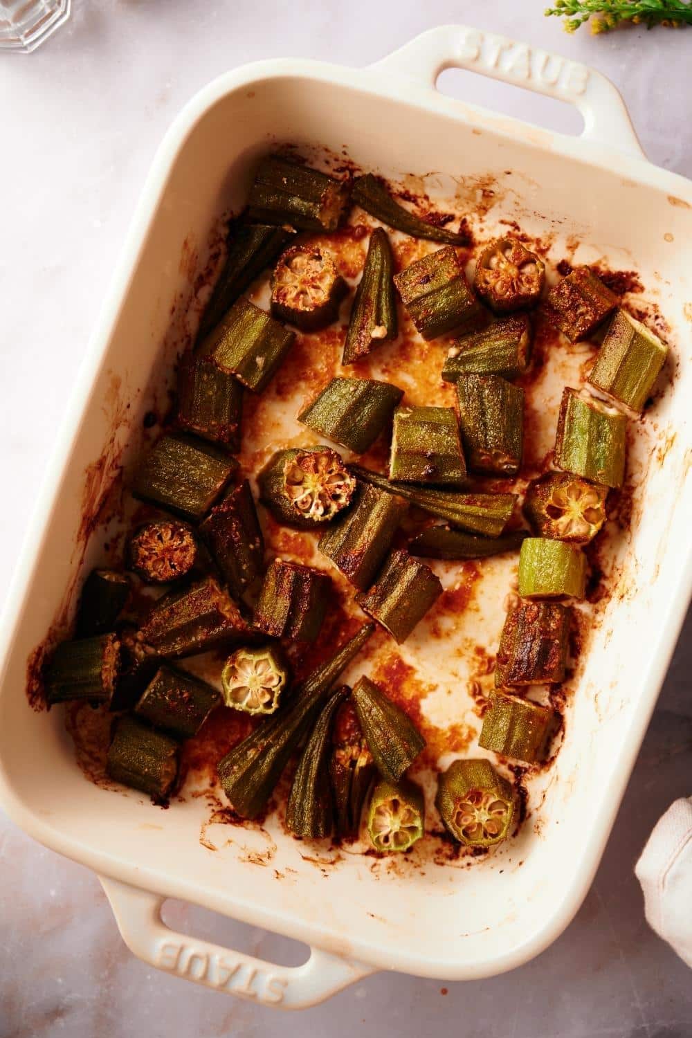 close up of cooked roasted okra in a white baking dish on a white marble countertop.