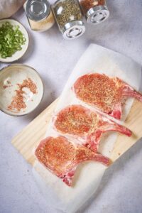 Three seasoned raw pork chops on a wooden cutting board with a sheet of parchment paper on top. Next to the board is an almost empty plate of spice rub, a small plate of chopped green onions, and three spice jars of garlic powder, paprika, and pepper.