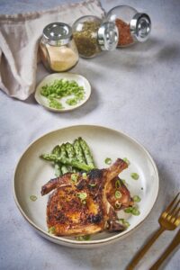 Two seared thin pork chops stacked in an ecru plate on top of 6 pieces of cooked aspargus. Next to the plate is part of a gold fork and knife and behind the plate are three small spice jars and a small plate of chopped green onions.