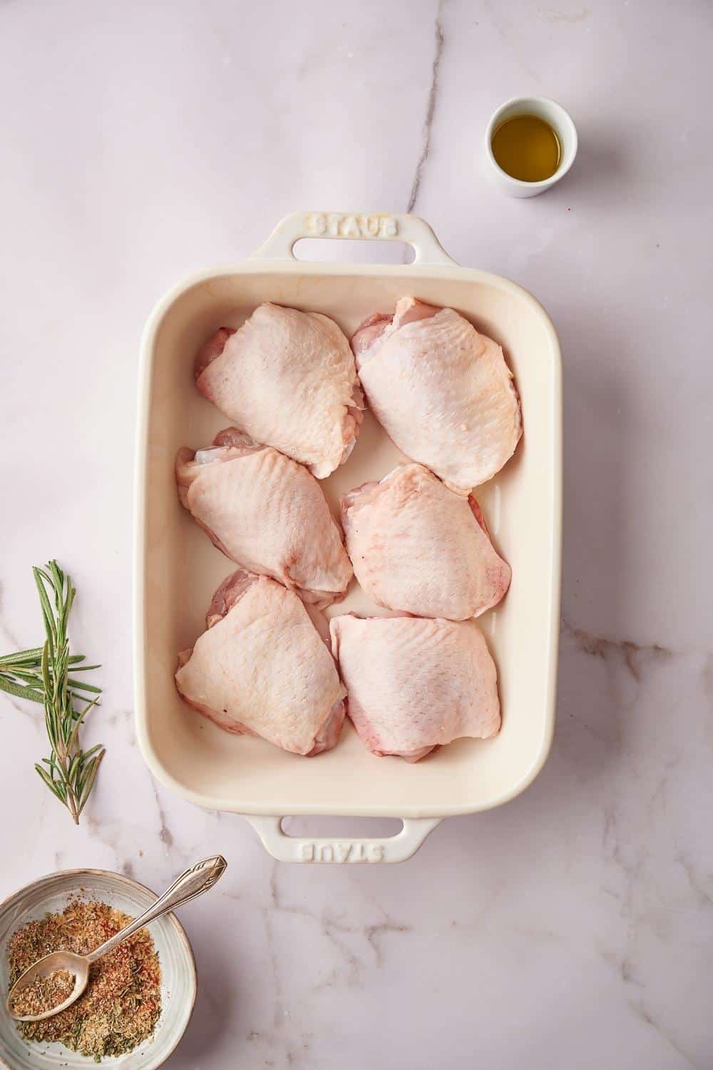 a baking tray with 6 raw chicken thighs in one layer. To the side of the tray is a small bowl of olive oil, a bowl of seasoning mix with a spoon laying on top, and a couple sprigs of fresh rosemary