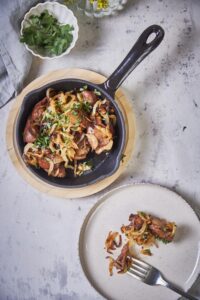 A small black serving pan filled with sauteed chicken livers and onions on a wooden plate. Next to the pan is a white plate with a serving of chicken livers and onions and a fork.