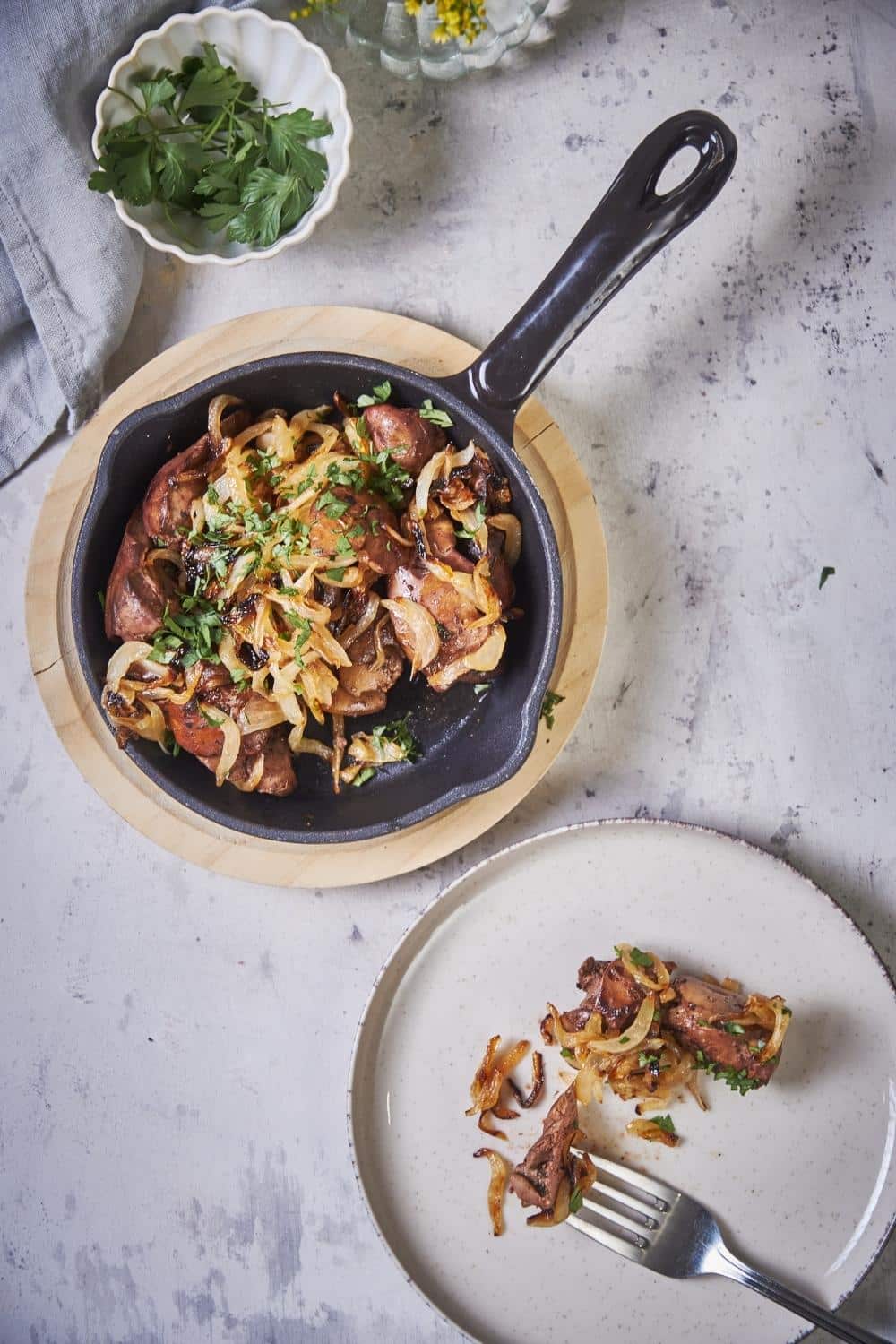 A small black serving pan filled with sauteed chicken livers and onions on a wooden plate. Next to the pan is a white plate with a serving of chicken livers and onions and a fork.