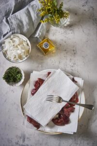 Raw chicken livers on a kitchen towel lined plate with another kitchen towel placed on top of them. A fork sits on the kitchen towel. To the side there's a bottle of olive oil, a bowl of sliced raw onions, and a smaller bowl of parsley.