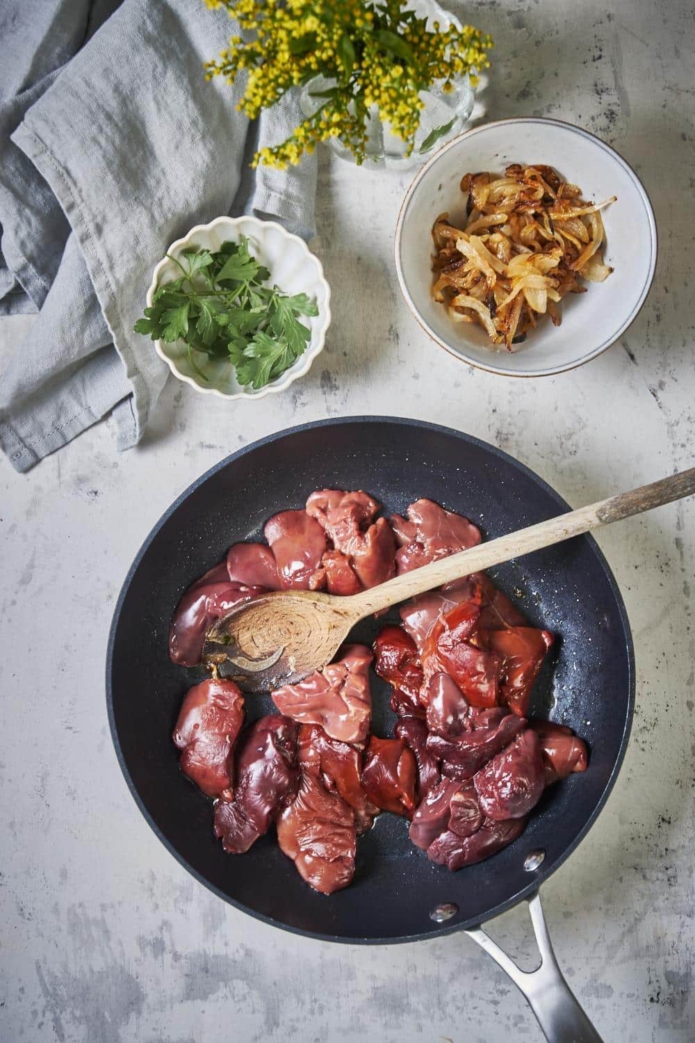 Raw chicken livers being cooked in a pan with a wooden spoon. Next to the pan is a bowl of cooked sliced onions and a smaller bowl of garnish.