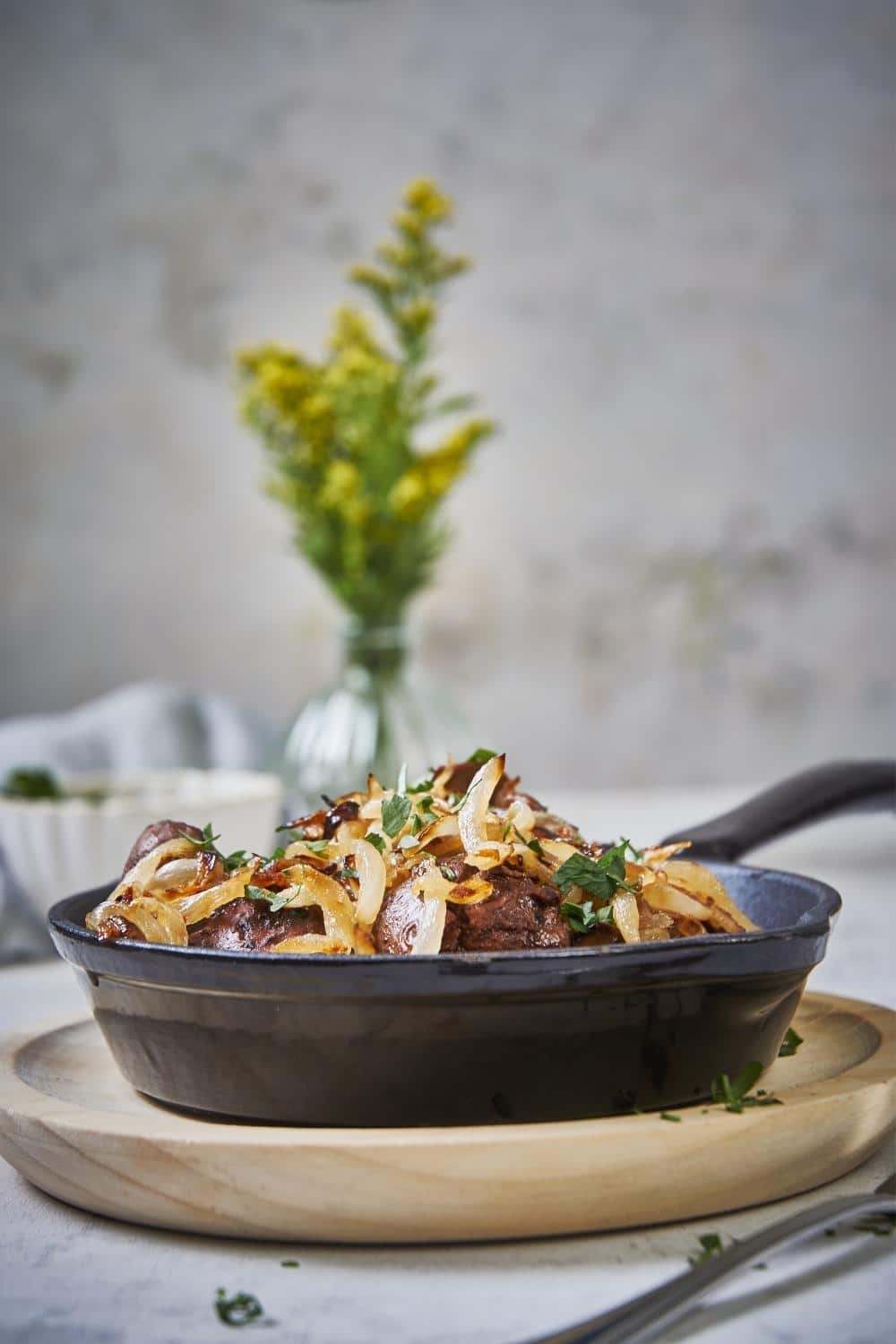 Chicken livers and onions garnished with parsley in a black pan on top of a wooden plate.