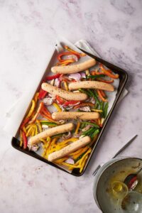 Uncooked chicken sausages, bell peppers, and onions coated with herbs and oil on a parchment paper lined baking tray.