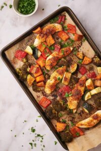 Baked chicken tenderloins and veggies in a parchment paper lined baking sheet. Part of a small bowl of herbs can be seen on the side. The chicken and veggies are garnished with chopped herbs, some of which are scattered lightly on the tabletop.