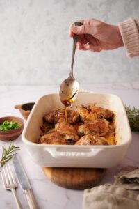 A hand holding a spoon dripping golden cooking juices over the baked chicken thighs.