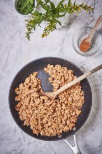 A skillet filled with cooked chorizo. A wooden spoon is scooping up some of the chorizo. Nearby is a smaller wooden spoon resting on a small glass bowl and a small ramekin filled with chopped cilantro.