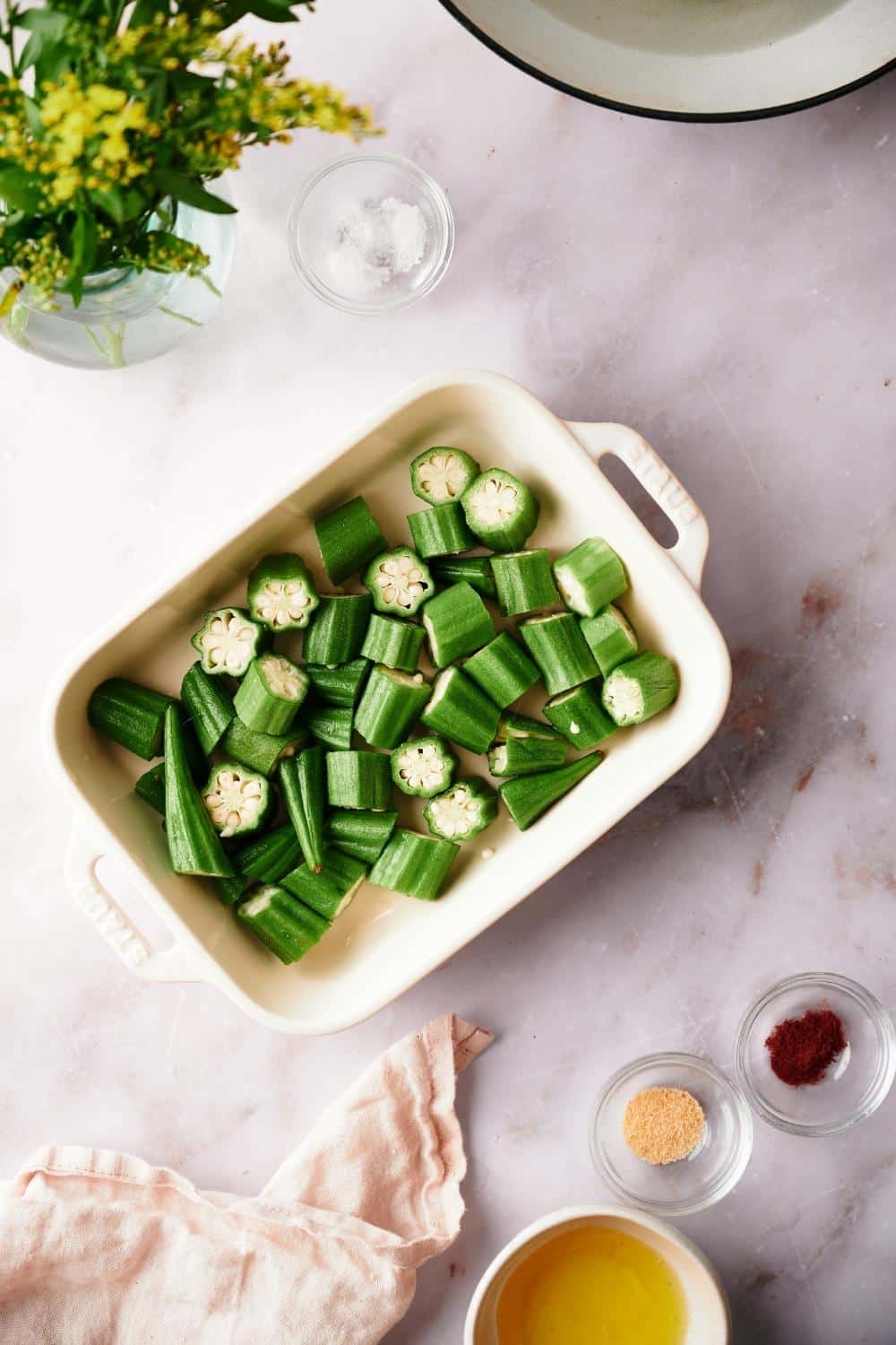 chopped okra pieces in a white baking dish on a marble countertop. Small bowls of garlic powder, paprika, salt, and melted butter surround the baking dish