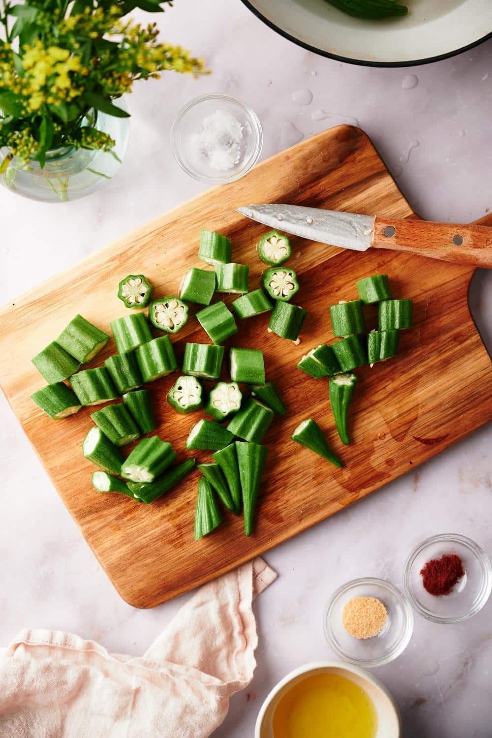 freshly chopped okra pieces on a cutting board on a marble countertop. a small knife with a wooden handle is on the side. Small bowls of garlic powder, salt, paprika, and melted butter surround the board as well as a small tea towel.