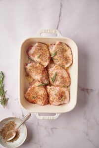 A white baking tray with 6 raw chicken thighs seasoned with spices and fresh rosemary. A single sprig of fresh rosemary sits to the side next to a small, half empty bowl of seasoning mix