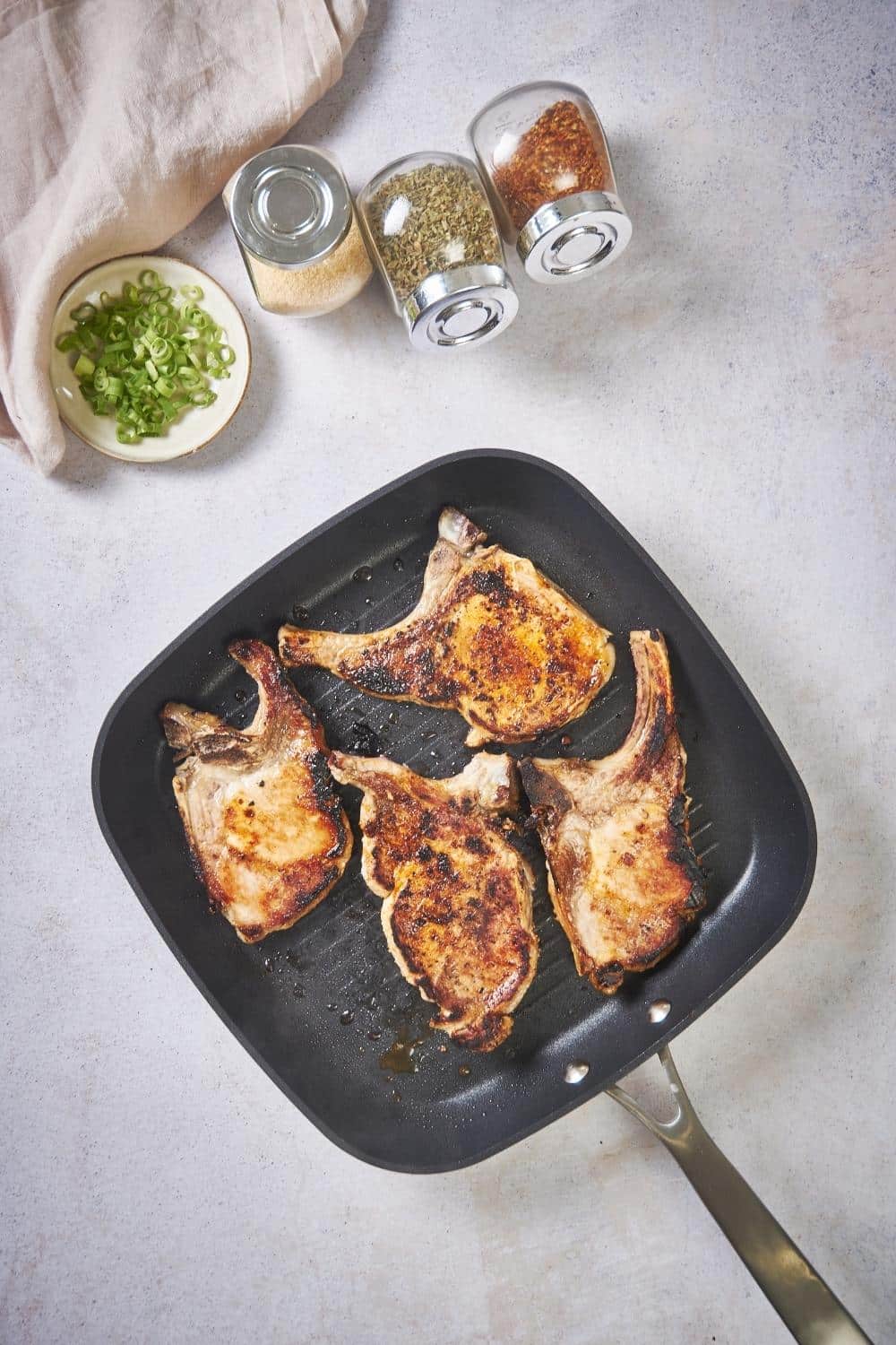 Four seared pork chops in a square grill pan. On the corner are three spice jars of garlic powder, paprika, and pepper, and a small plate of chopped green onions.