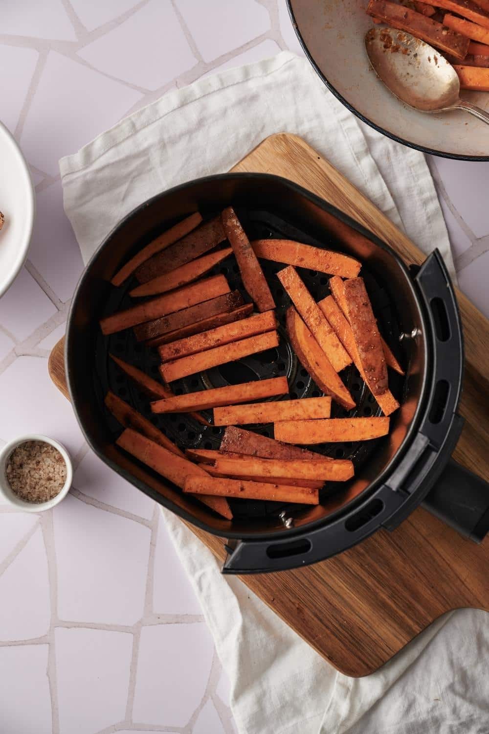 Seasoned raw sweet potato fries arranged in one layer in an air fryer basket resting on top of a wooden cutting board. A bowl with more air fryer fries can be seen on the upper corner.