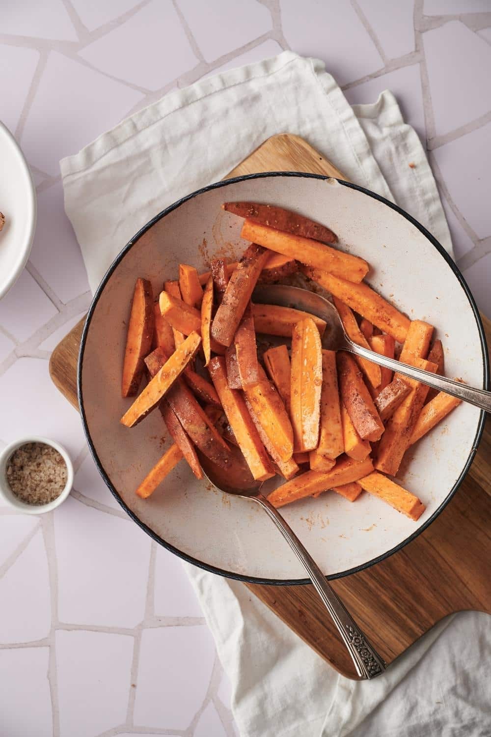 Two spoons tossing raw sweet potato fries and seasoning together in a large white bowl on a cutting board.