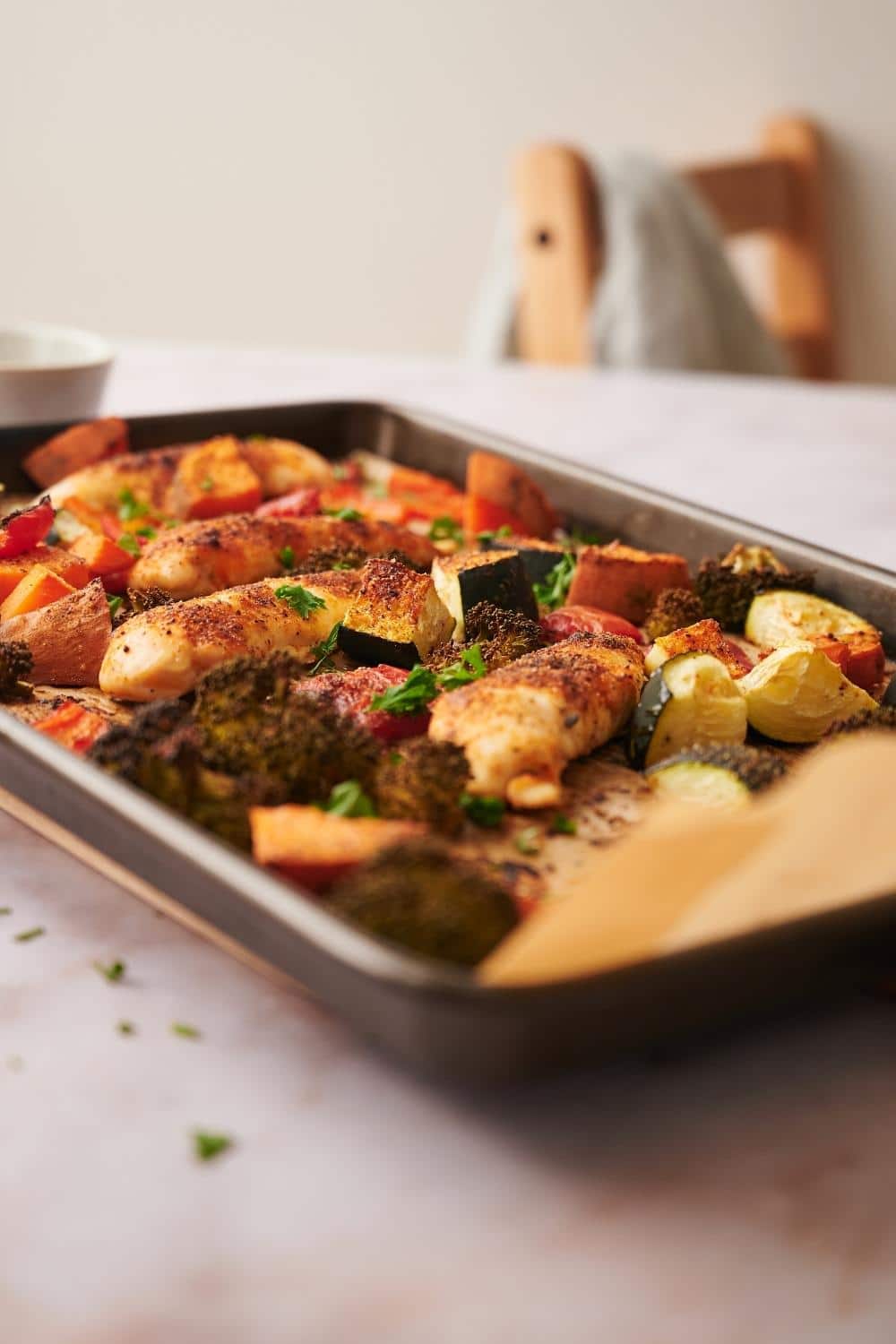 Chicken tenderloins and chopped veggies in a parchment paper lined baking sheet. It's garnished with herbs, a few of which are scattered on the table top. A small bowl can be seen in the back.