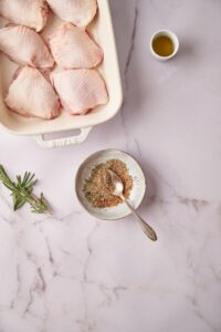 a small bowl of herbs and spices mix with a spoon. Surrounding it is a couple of sprigs of fresh rosemary, a small bowl of olive oil, and a baking tray with 6 raw chicken thighs.