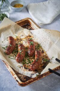 Four roasted brats arranged neatly on a baking sheet lined with parchment paper. The brats are garnished with fresh parsley. A pair of black tongs sits on the corner of the sheet and small bowls of mustard and parsley can be seen in the back.