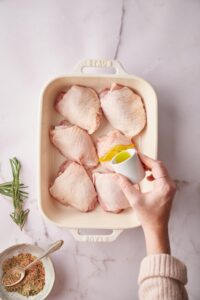A hand pouring olive oil over raw chicken thighs in a baking dish.