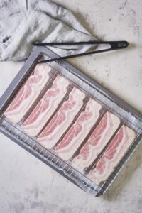 6 pieces of uncooked turkey bacon arranged neatly on a baking rack on top of a baking sheet lined with parchment paper. A pair of black rubber tongs sits on the edge of the baking sheet on top of a tea towel.