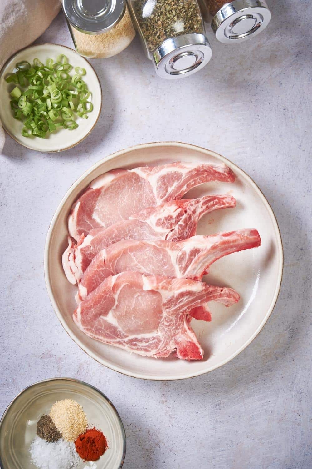 Four raw pork chops on a plate. A small plate of unmixed spices can be seen on the side, and on the other side there are three spice jars of garlic powder, paprika, and pepper, and a small plate of chopped green onions.
