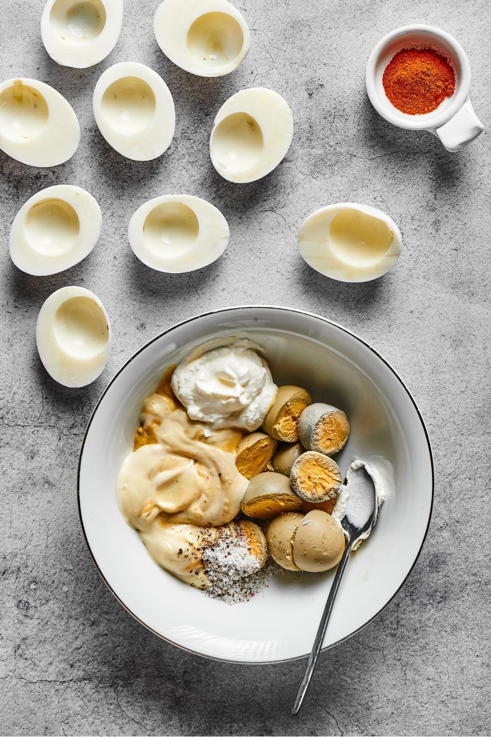 A bowl of hard boiled egg ingredients. Behind it are nine hard boiled egg whites.