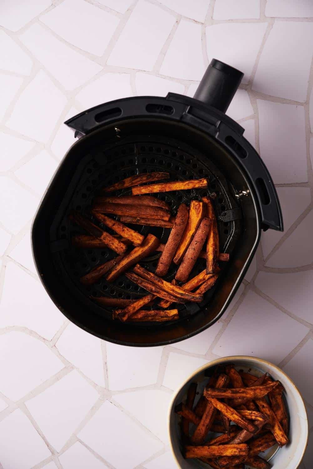 Cooked sweet potato fries in an air fryer basket resting on a countertop. The bottom right side is a small bowl of cooked sweet potato fries from the first batch.
