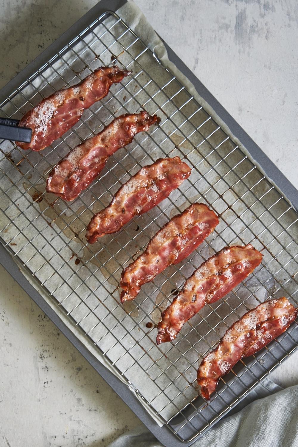 6 pieces of baked turkey bacon slices on a baking rack. A pair of black tongs is flipping the rightmost piece of bacon.