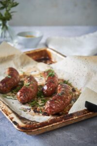 Four roasted brats garnished with chopped fresh parsley. They're arranged on a parchment paper lined baking sheet and pair of black tongs sits on the corner. A small bowl of mustard can be seen in the background next to a white tea towel.