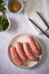 Four uncooked brats on a white plate. A pair of black tongs sits on a tea towel next to the plate. Next to it is a small bowl of mustard and a small bowl of parsley.