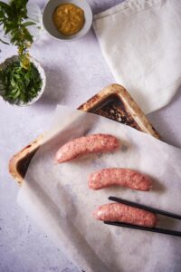 Three raw brats on a parchment paper lined baking sheet. A pair of black tongs is placing the third brat. A tea towel, a small bowl of mustard, and a small bowl of fresh parsley can be seen on the side.