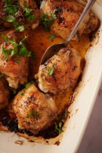 Close up of a spoon taking out a baked chicken thigh coated in seasonings and golden cooking juices from a tray of baked chicken thighs.