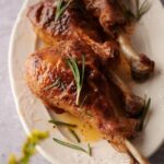 Three roasted turkey legs on an oval embossed plate garnished with rosemary leaves and cooking juices. A few small yellow flower buds can be seen in the foreground.