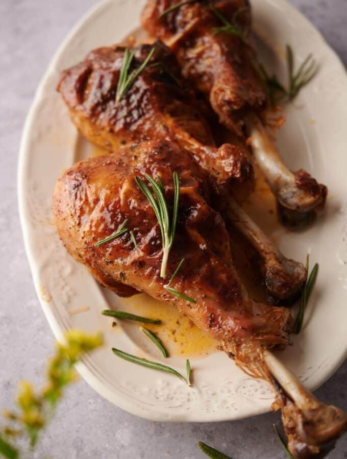 Three roasted turkey legs on an oval embossed plate garnished with rosemary leaves and cooking juices. A few small yellow flower buds can be seen in the foreground.