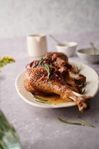 Close up of roasted turkey legs on an oval plate garnished with rosemary leaves and cooking juices. Two small bowls with metal spoons can be seen in the background next to a small sauce pitcher.