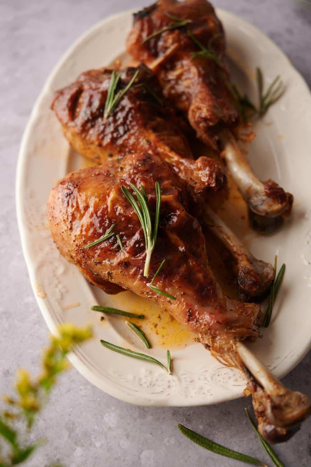 Three roasted turkey legs on an oval embossed plate garnished with rosemary leaves and cooking juices. A few small yellow flower buds can be seen in the foreground.
