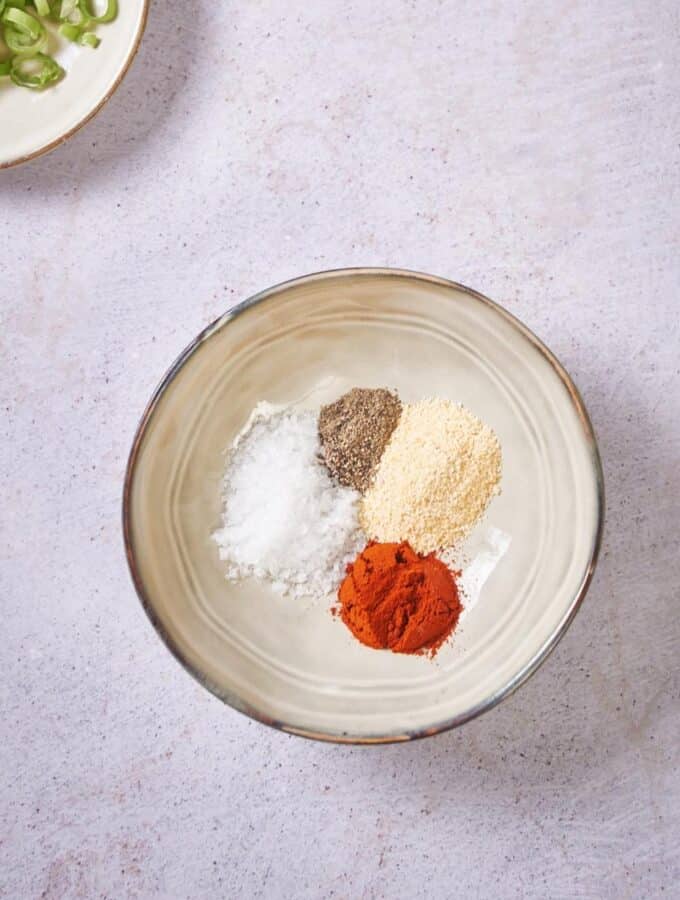 Paprika, garlic powder, pepper, and salt in a small plate. Part of a set of spice jars and a small plate of chopped green onions can be seen on the side.