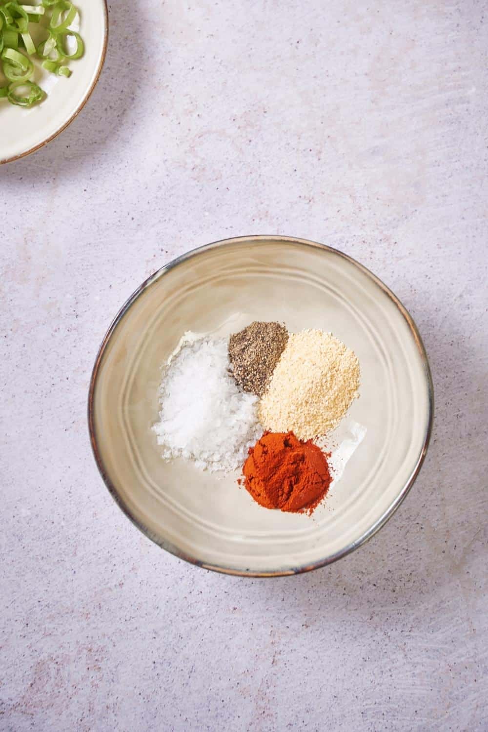 Paprika, garlic powder, pepper, and salt in a small plate. Part of a set of spice jars and a small plate of chopped green onions can be seen on the side.