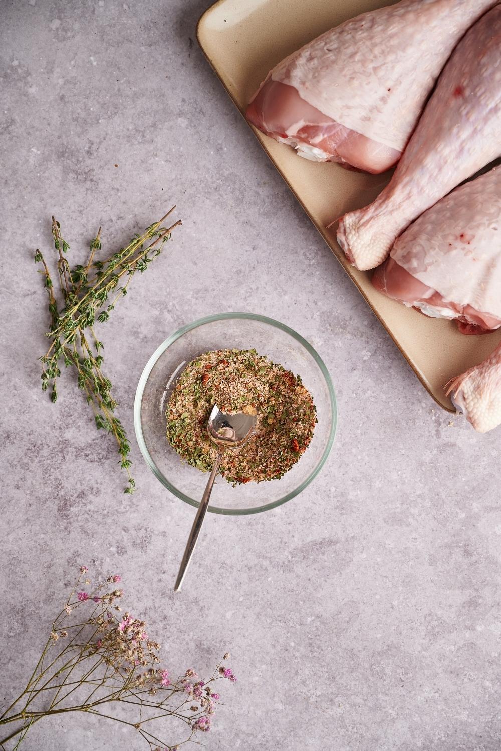 An herb and spice mix in a small glass bowl with a metal spoon. Next to the bowl is some fresh thyme. Part of a plate of raw turkey legs can be seen on one side and dried purple flowers are placed on the other side.