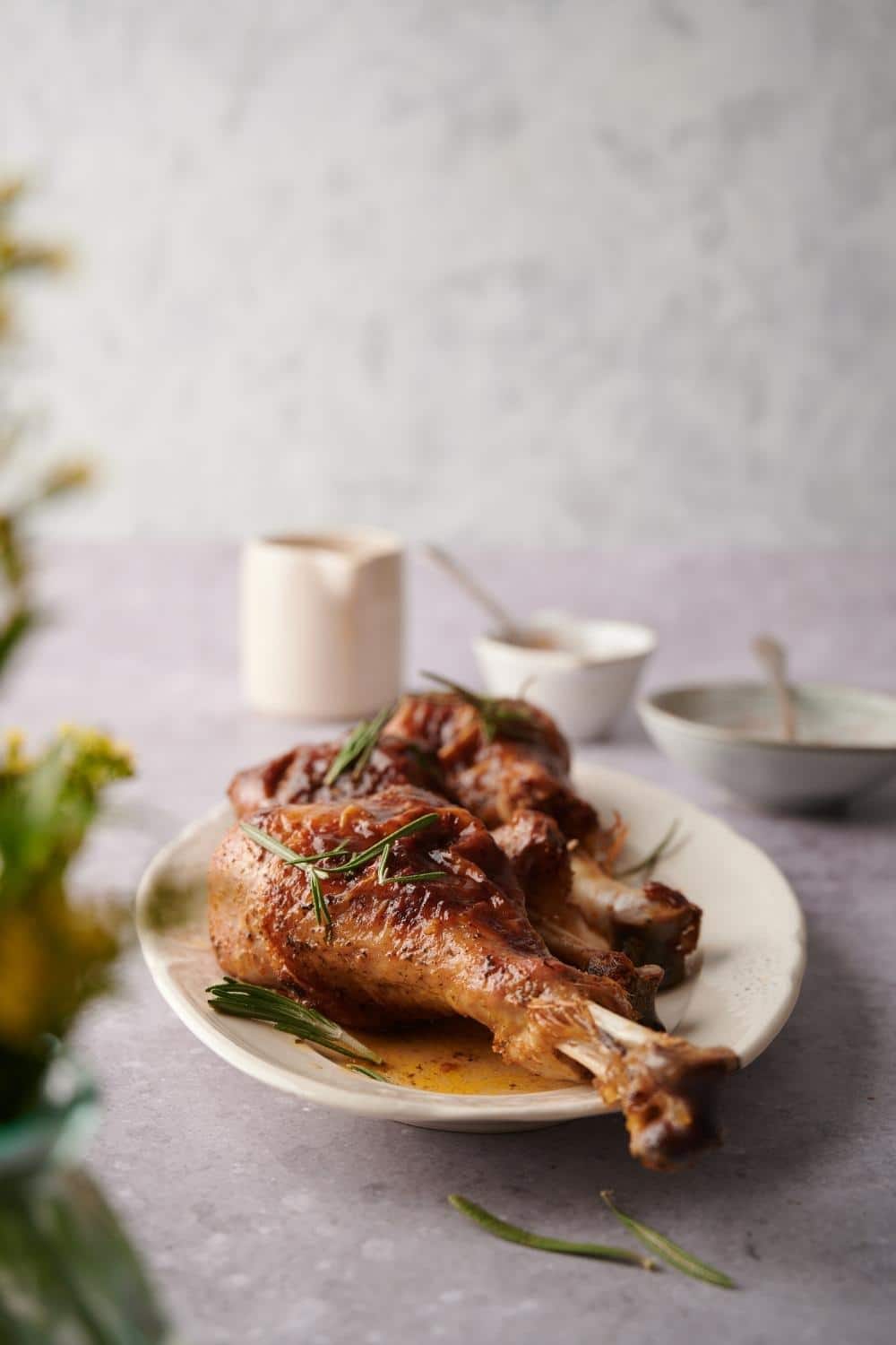 Three roasted turkey legs on an oval plate garnished with rosemary leaves and cooking juices. A small sauce pitcher and two small bowls with metal spoons can be seen in the background.