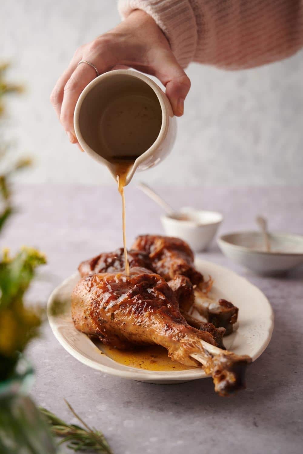 A small ceramic pitcher of golden cooking juices being poured over roasted turkey legs on an oval plate.