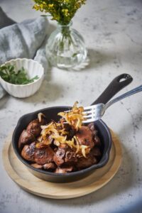 A fork placing cooked onions over sauteed chicken livers in a small black serving pan. The pan is on a wooden plate and there's a bowl of parsley in the back.