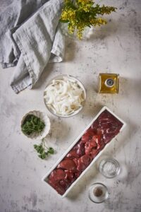 A narrow rectangular bowl filled with raw chicken livers, a bowl of sliced raw onion, a small bowl of parsley, a bottle of olive oil, and two small glass bowls holding salt and pepper.