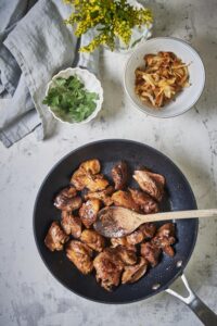 Golden brown sauteed chicken livers in a skillet with a wooden spoon. Next to the pan is a bowl of cooked onions and a smaller bowl of garnish.