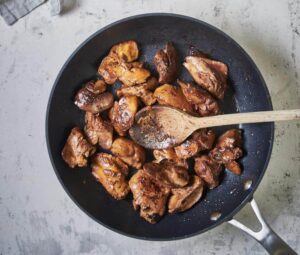 Golden brown sauteed chicken livers in a skillet with a wooden spoon. Next to the pan is a bowl of cooked onions and a smaller bowl of garnish.