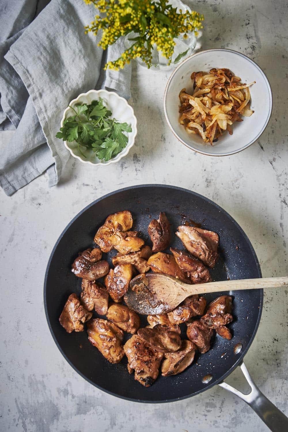 Golden brown sauteed chicken livers in a skillet with a wooden spoon. Next to the pan is a bowl of cooked onions and a smaller bowl of garnish.