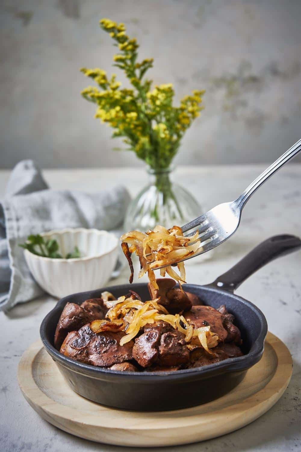 A fork gently placing cooked onions on top of sauteed chicken livers in a small black serving pan.
