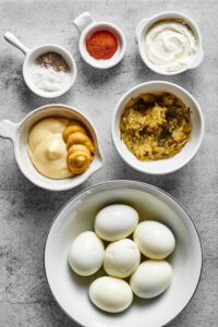 A bowl of six hard boiled eggs, behind it is a bowl of sweet relish, a bowl of mayo and mustard, a bowl of salt and pepper, a bowl of cayenne pepper, and a bowl of cream cheese.
