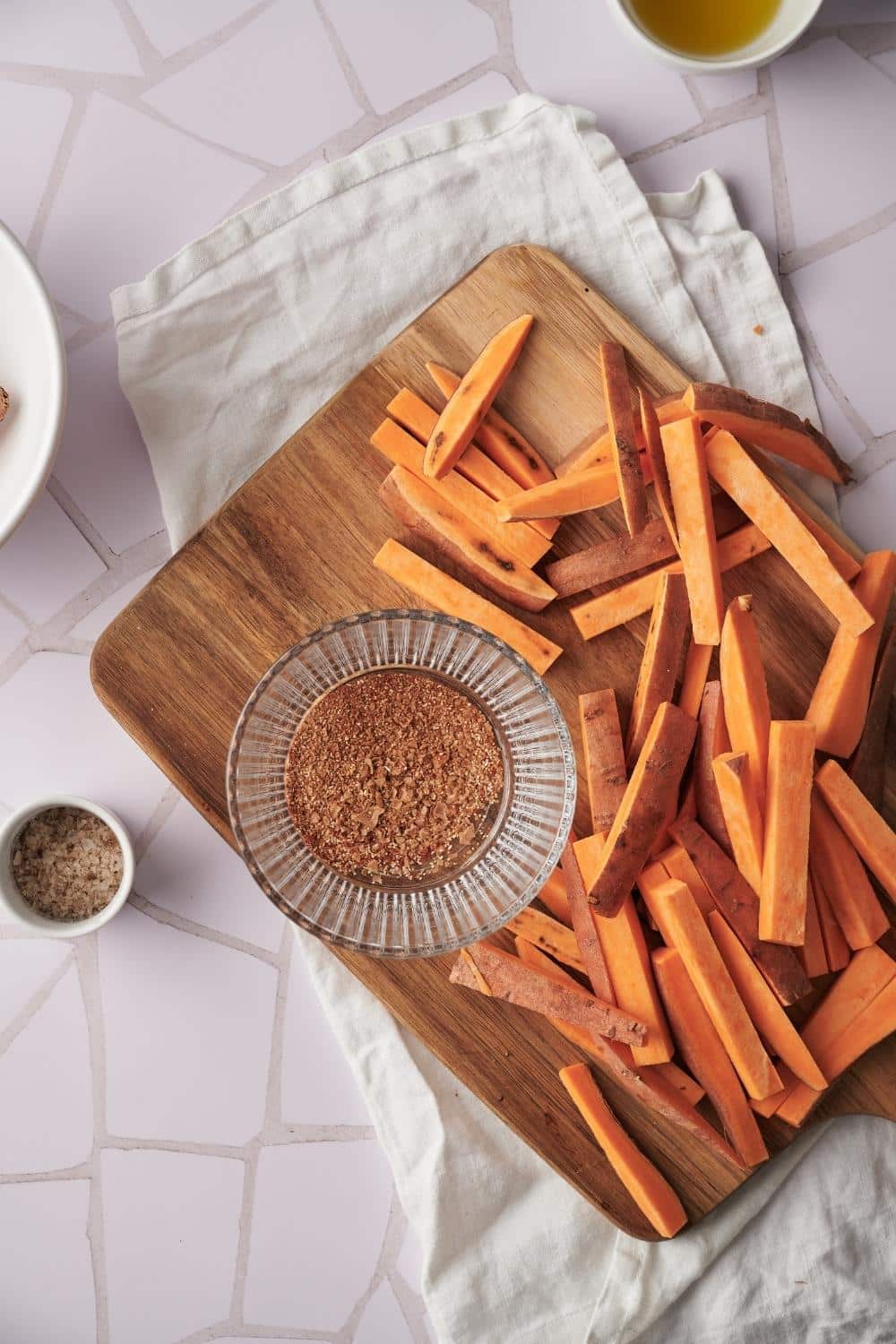 chopped sweet potato fries on a wooden cutting board resting on a tea towel. A glass bowl of seasoning mix sits next to the fries.